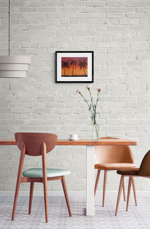 Image of palm tree oil painting in a black frame hanging on a brick wall. A table with a cup of coffee on it and a vase with flowers. 
